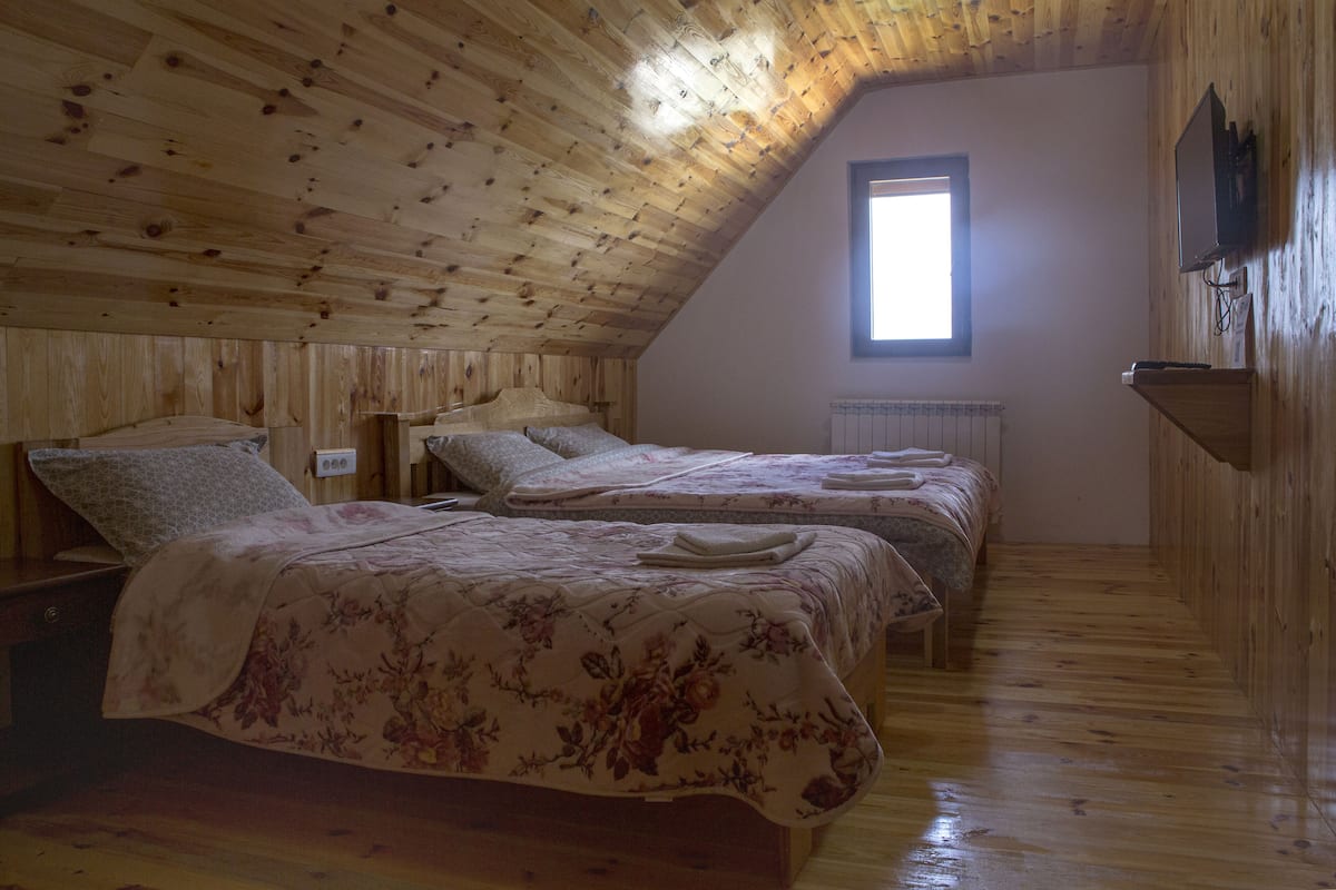 A cozy attic bedroom with wooden walls and a sloped ceiling, featuring two beds with floral bedding and a small window providing natural light.