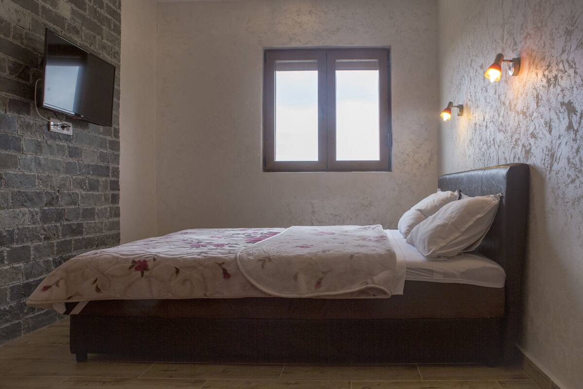 A bedroom with a bed, a TV mounted on a brick wall, and a window with two light fixtures.