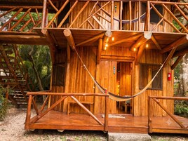 Una cabaña de madera con una hamaca, escaleras de madera y una puerta que da al interior.