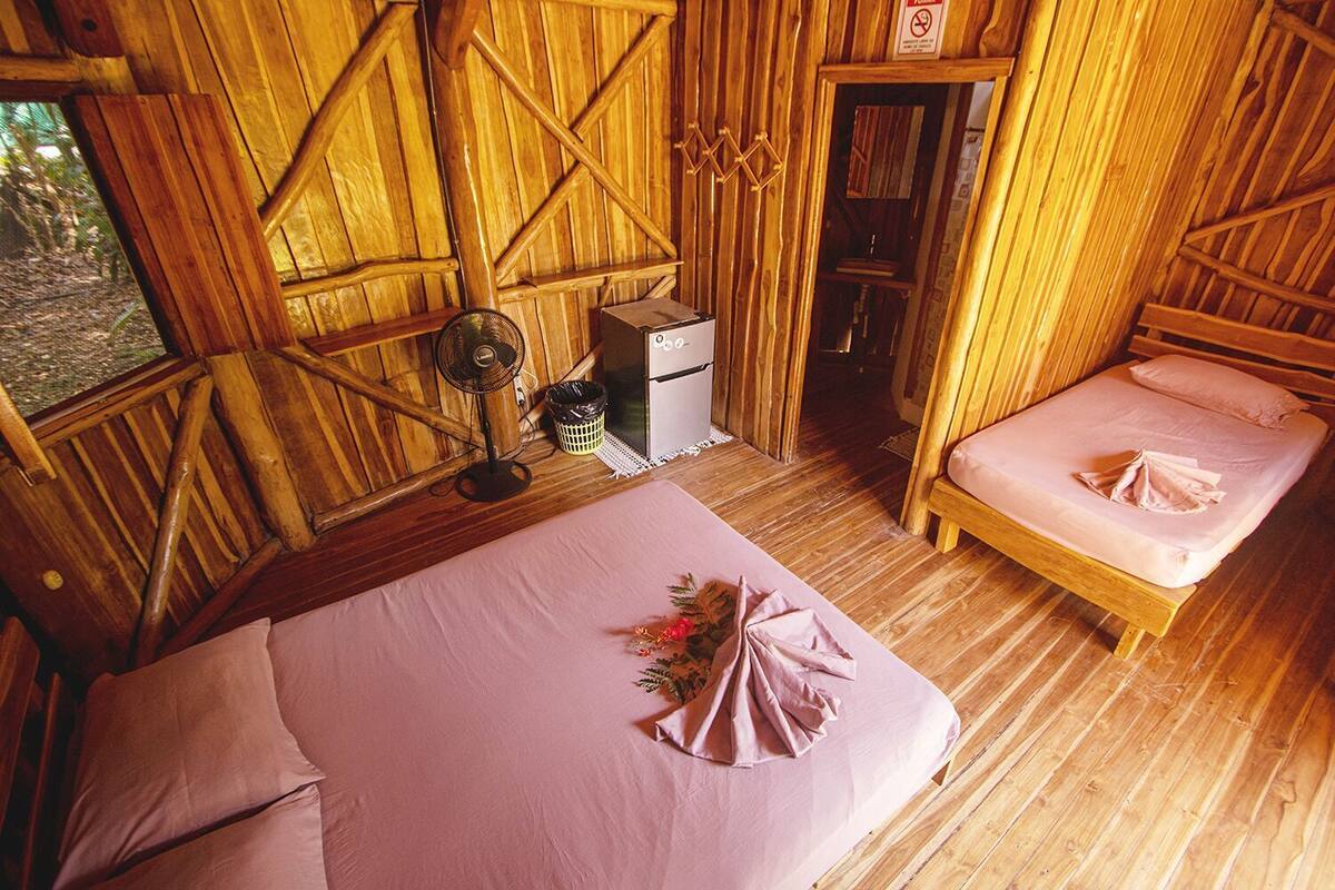 Una cabaña de madera con una cama, un escritorio pequeño, un ventilador y un cubo de basura.