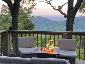 Une terrasse dotée de deux fauteuils confortables, disposés face à un foyer extérieur, le tout entouré d’arbres et offrant une vue sur des collines lointaines.