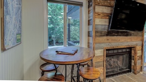 Un coin repas avec une table ronde et des tabourets de bar, une cheminée et une grande fenêtre donnant sur les arbres.