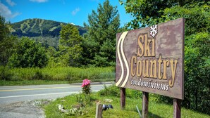 Un panneau en bois portant l’inscription « Ski Country condominiums », situé dans un cadre rural, avec une route et de la végétation.