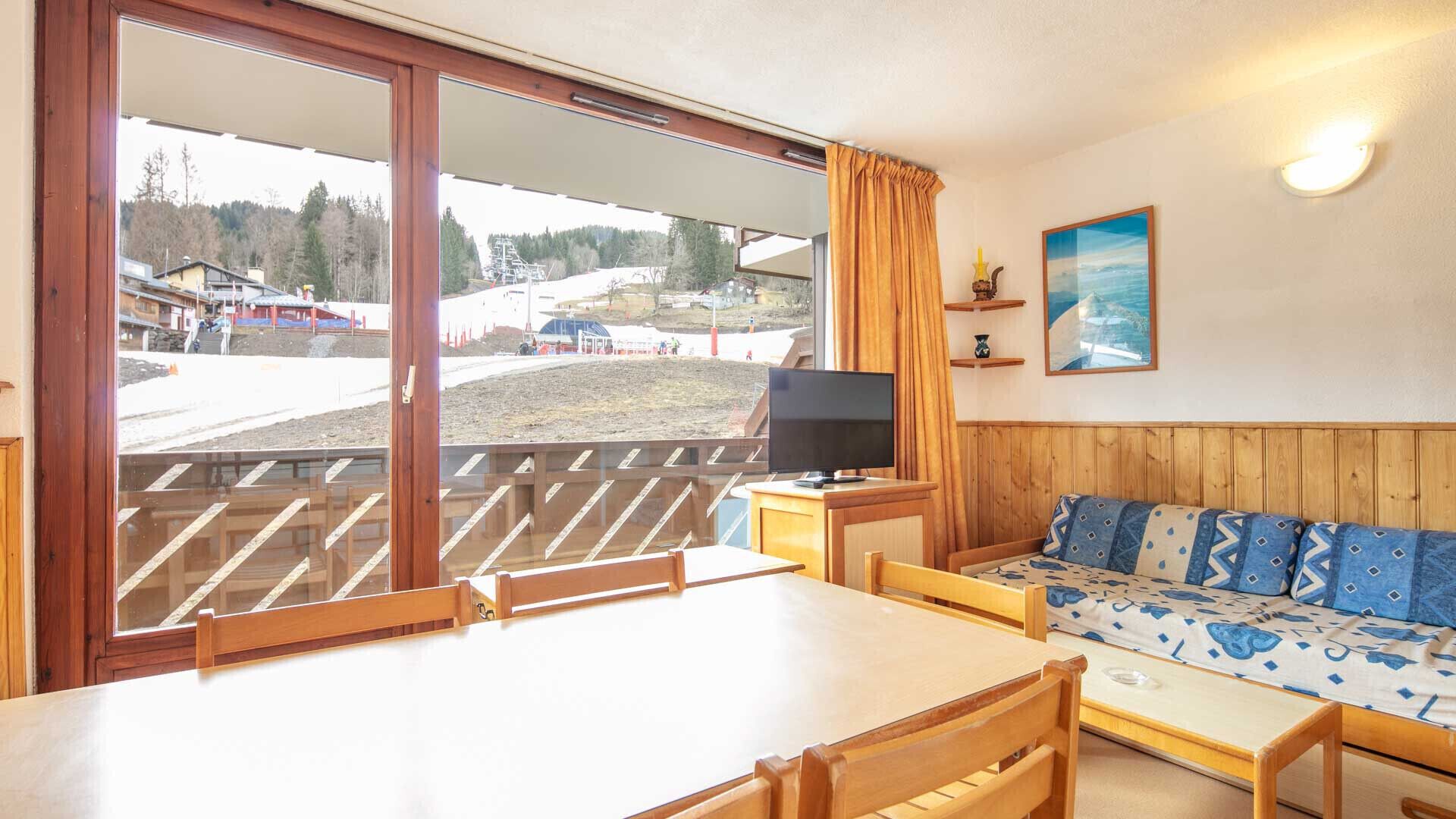 Ein Zimmer mit einem Holztisch, einer Couch mit gemusterten Kissen, einem Fernseher und einem großen Fenster mit Blick auf eine Skipiste.