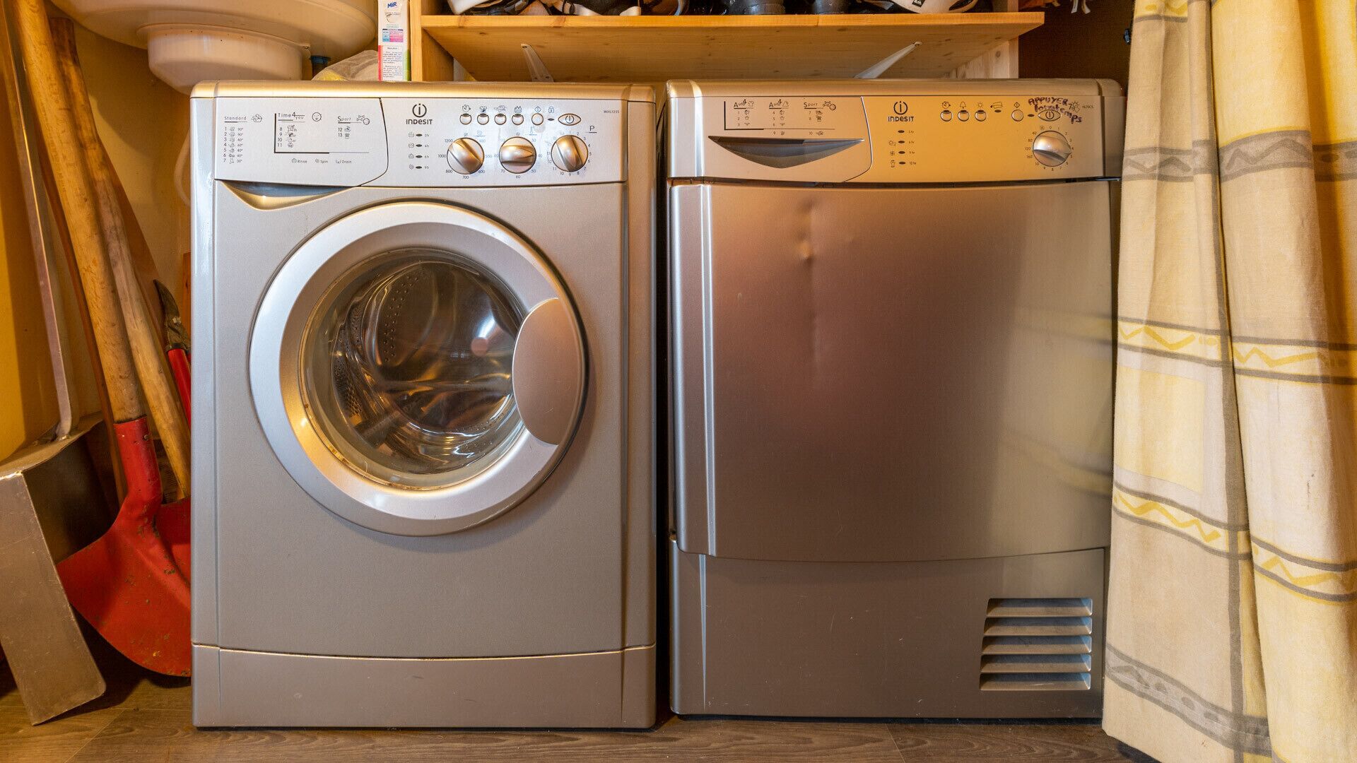 Two stacked washing machines in a laundry room with a wooden shelf above and a curtain on the right.