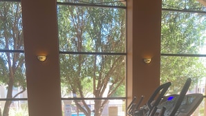 Ett gym med löpband och crosstrainers, stora fönster med utsikt över träd och en byggnad utanför.