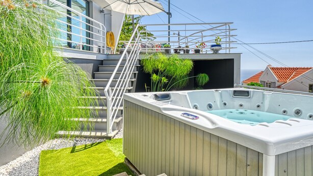 Ein moderner Außenbereich mit Whirlpool, eine Leiter führt zu einer Terrasse, umgeben von üppiger Vegetation und blauem Himmel.