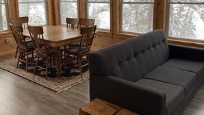 Ein gemütliches Blockhaus-Interieur mit Esstisch, Stühlen, einer Couch und großen Fenstern mit Blick auf schneebedeckte Bäume.