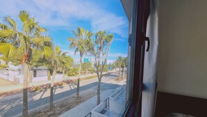 A view from a window with a concrete planter filled with white pebbles, a street with palm trees, and a clear blue sky. - STAY Anemone 20 (Protaras)