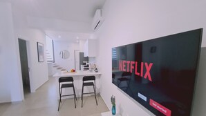 A modern living room with a large flat-screen TV displaying the Netflix logo, a kitchen area in the background, and a dining space with two black chairs. - STAY Anemone 20 (Protaras)