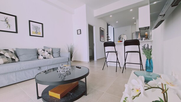 A modern living room with a sofa, a coffee table, and two chairs. There is a kitchen area in the background with a counter and cabinets. - STAY Anemone 20 (Protaras)