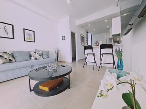 A modern living room with a sofa, a coffee table, and two chairs. There is a kitchen area in the background with a counter and cabinets. - STAY Anemone 20 (Protaras)