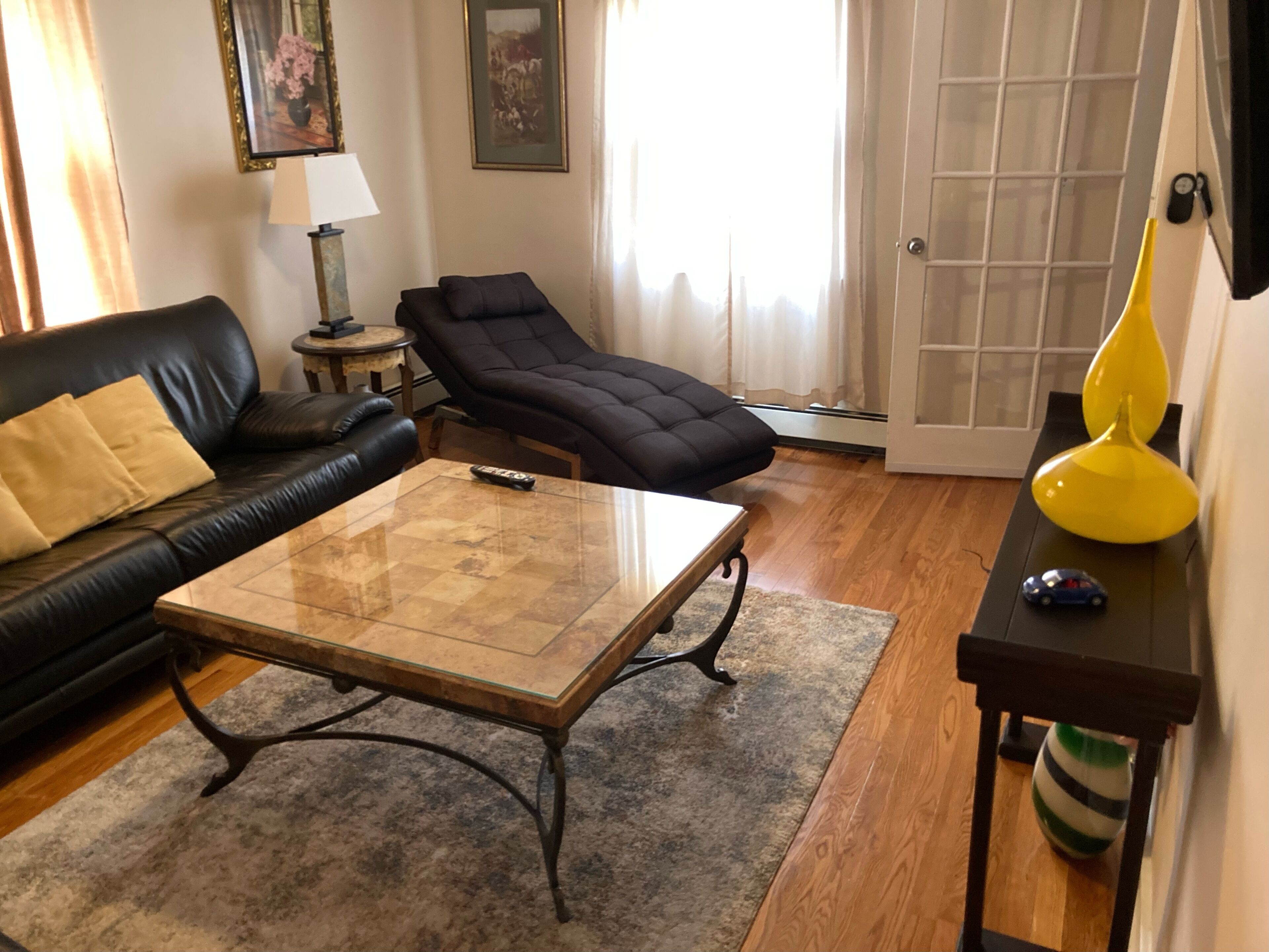 A living room with a black leather sofa, a black recliner, a glass coffee table, a lamp, and a small yellow vase.