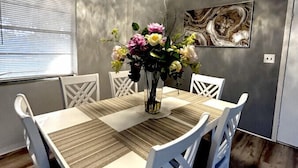 A dining area with a white table and chairs, a vase of flowers, a ceiling light, and a painting on the wall. - Beach Break House by the Surf (Daytona Beach)