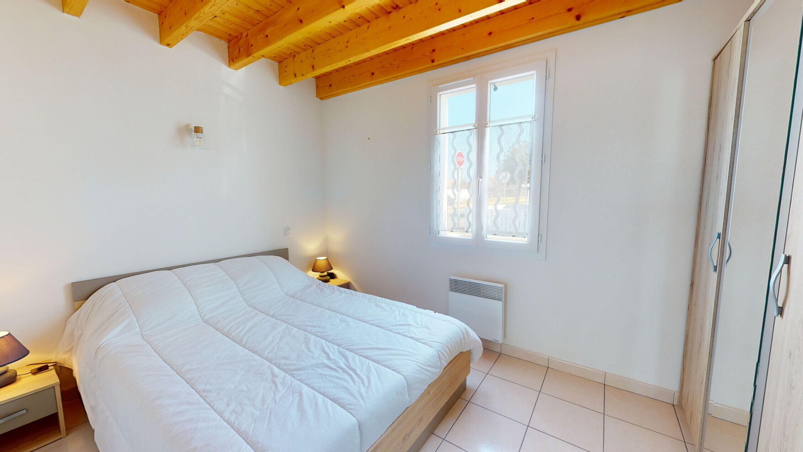 Un dormitorio con techo de madera, una cama, una mesita de noche con lámpara, una ventana y un radiador.