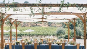 Un área de comedor al aire libre con mesas dispuestas para comensales, rodeada por una estructura de pérgola de madera.