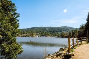 A lakeside view with a wooden fence, rocky shoreline, and a forested hill in the background. - Modern Crestline Cabin Near Creek & Lake Gregory (Crestline)
