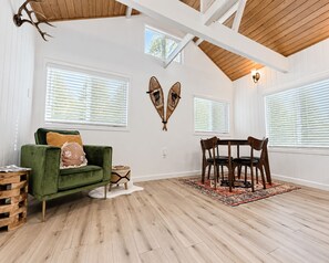 A room with a green sofa, wooden dining table, and chairs, white walls, and wooden beams on the ceiling. - Modern Crestline Cabin Near Creek & Lake Gregory (Crestline)
