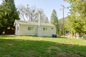 Une petite maison de plain-pied, dotée d’un toit à pignon et d’une cheminée. Elle est entourée d’une cour verdoyante, avec des arbres et une clôture en bois.