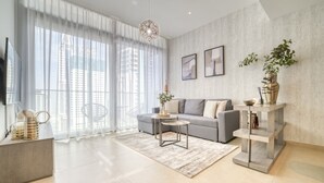 A modern living room with a grey sofa, a coffee table, and a TV on a stand. There is a large window with sheer curtains, a plant on a side table, and a decorative light fixture.