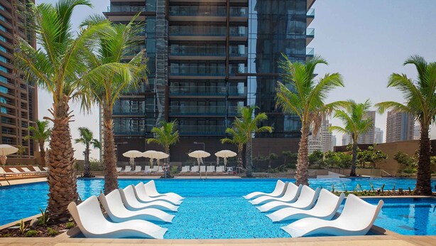A modern hotel pool with white lounge chairs, palm trees, and a high-rise building in the background.