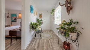 Un pasillo con suelo de madera, una cama con cabecera blanca, un escritorio con lámpara y plantas en macetas sobre estanterías.