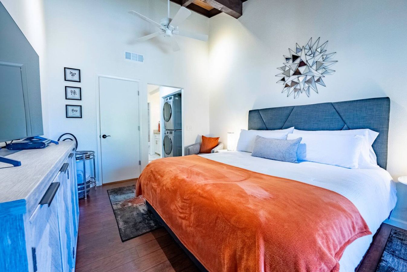 Ein modernes Hotelzimmer mit einem großen Bett, einem Deckenventilator, einem an der Wand befestigten abstrakten Kunstwerk und einem sichtbaren Waschbereich.