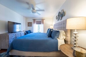 A bedroom with a large bed, a television on a wooden cabinet, a ceiling fan, and a window with curtains.