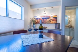 Un comedor con una mesa de madera, tres copas de vino y una botella. Hay un cuadro en la pared detrás de la mesa y un jarrón con plantas secas en la encimera.