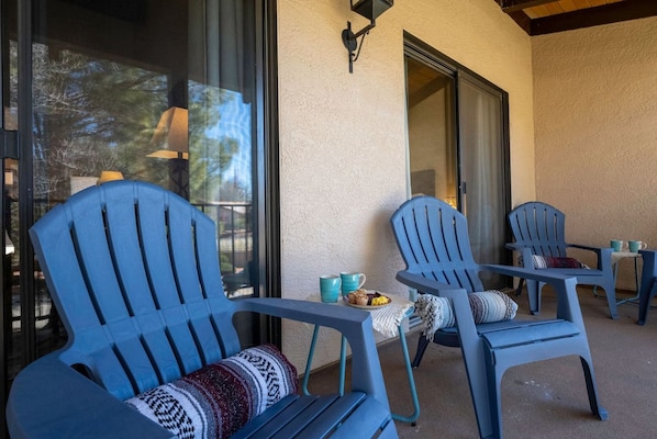 A patio area with blue Adirondack chairs, a small table, and a window with a view of trees.
