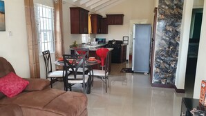 A spacious living room with a brown sofa and a dining area with red chairs. A kitchen with dark wooden cabinets and a blue refrigerator is visible in the background. - Cozy retreat pool access super close to the Town. Entire home available to you! (Old Harbour)