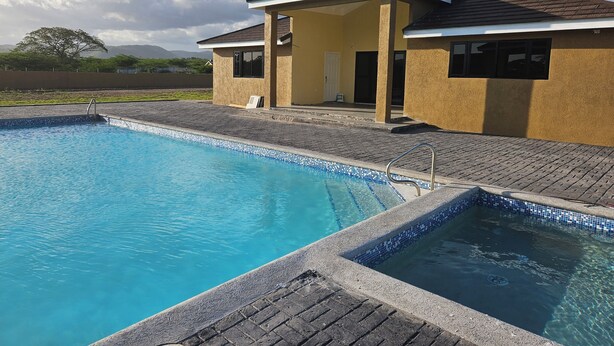A swimming pool with a tiled edge, a covered patio area, and a building with a beige exterior and a brown roof. - Cozy retreat pool access super close to the Town. Entire home available to you! (Old Harbour)