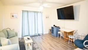 Una sala de estar con un sofá, una mesita, un televisor montado en la pared y un escritorio con sillas.