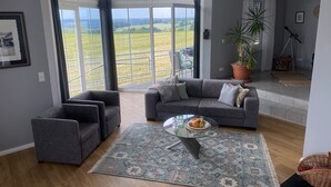 A modern living room with a grey sofa, a glass coffee table, and a large window offering a view of a green landscape. - Traumblick in die Natur (Hennef (Sieg))