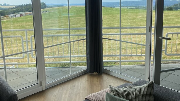 A corner room with large glass doors leading to a balcony, a grey sofa, and a view of a green field. - Traumblick in die Natur (Hennef (Sieg))