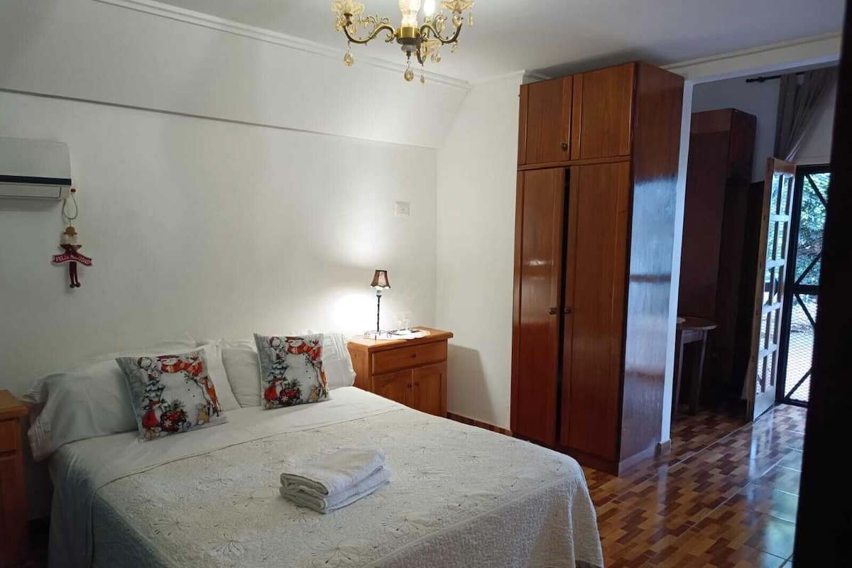 Een slaapkamer met een bed, nachtkastjes, een kroonluchter, een houten garderobe en een uitzicht door een deur.