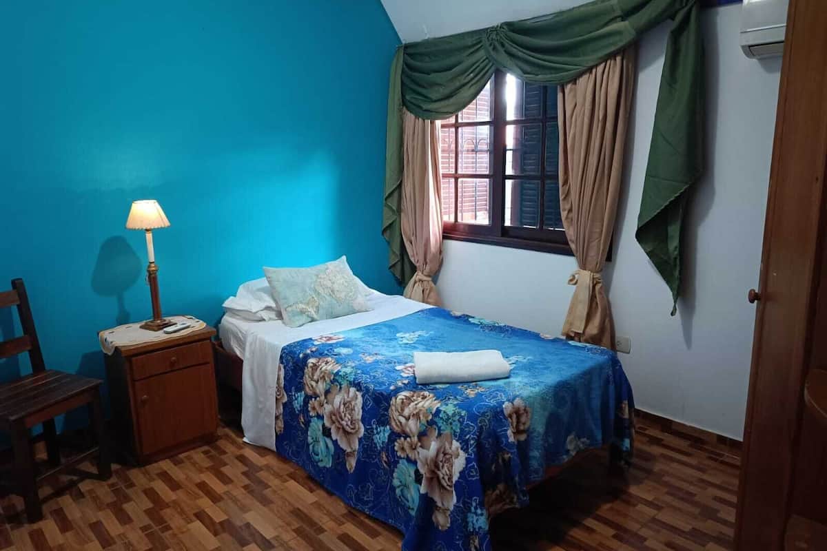 Een slaapkamer met een bed, nachtkastje, lamp, raam met gordijnen en een houten vloer.