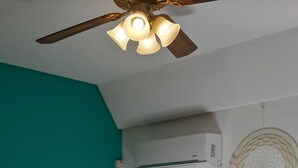 Een slaapkamer met een teal accentmuur, een plafondventilator, een airconditioning, een bedlampje, een bed met een geruite deken en een droomvanger aan de muur.