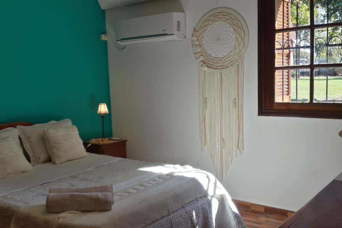 Een slaapkamer met een bed, een bedlamp, een wandairconditioner en een raam met uitzicht op groen.