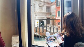 Une personne est assise près d’une fenêtre, en train de lire un magazine, et la fenêtre donne sur un bâtiment historique doté d’une grille en fer forgé.