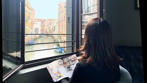 Une personne est assise près d’une fenêtre, un magazine à la main, et la vue donne sur un canal et des bâtiments.