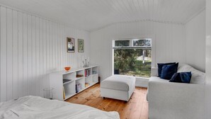 Ein Zimmer mit Holzboden, weißen Wänden, einem weißen Bett, einem weißen Sofa, einem Fenster mit Blick auf Grünflächen und einem Bücherregal mit Büchern und Dekoration.