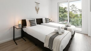 A modern bedroom with two beds, a bedside table with a lamp, a macrame wall hanging, and a large window with a view of greenery. - Sea & Sun Apartment - Pool with Sea View (Nazaré)