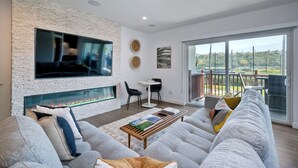 A modern living room with a fireplace, grey sofas, a coffee table with books, and a balcony with a view. - La Costa Luxury Condo | Fully Remodeled (Carlsbad)