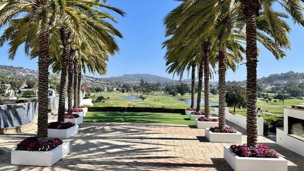 A paved pathway lined with palm trees and flower beds, leading to a golf course with a lake and houses in the background. - La Costa Luxury Condo | Fully Remodeled (Carlsbad)