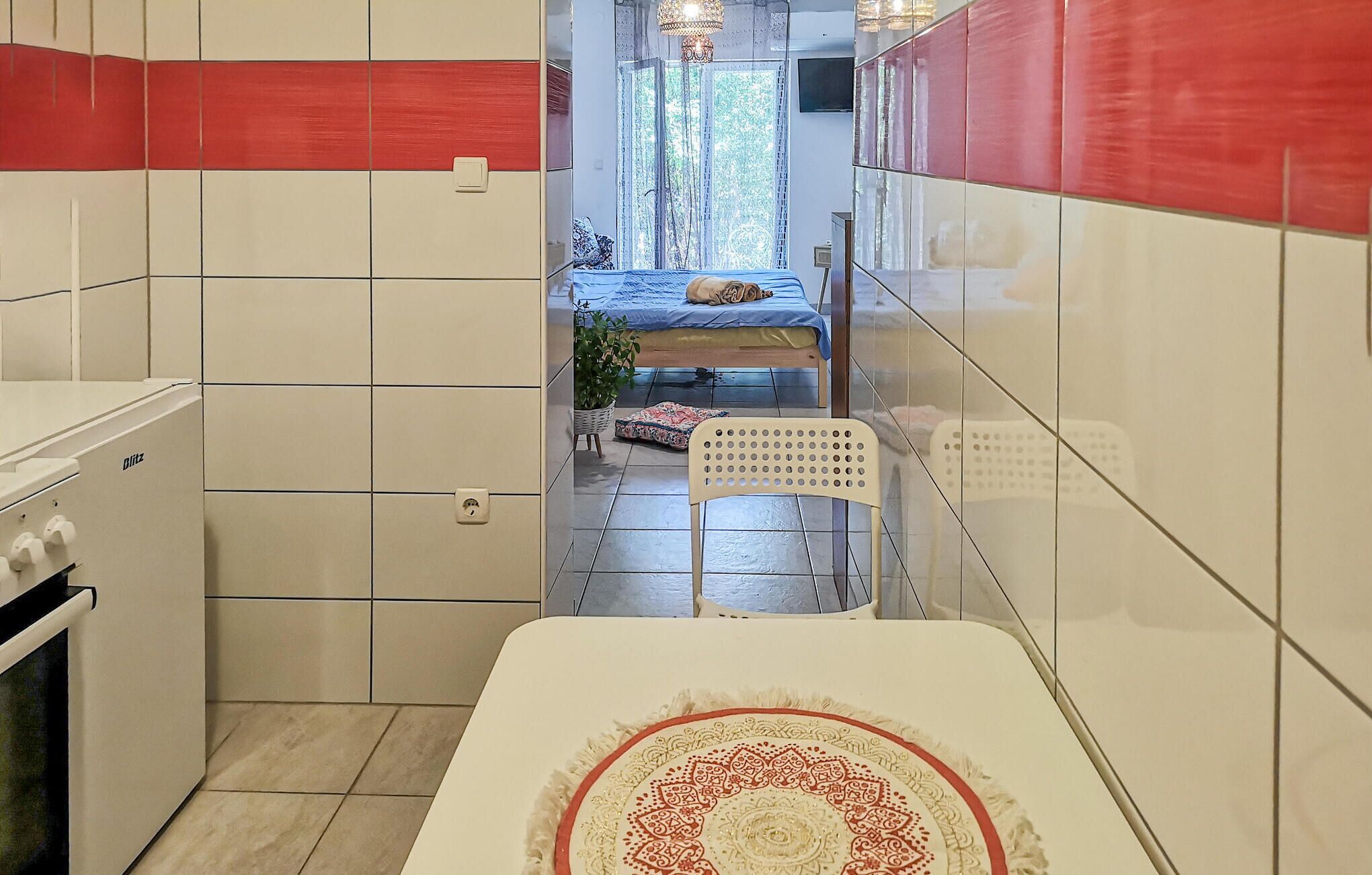 A narrow kitchen with white and red tiles, a stove, and a bed with a blue bedspread in the background.