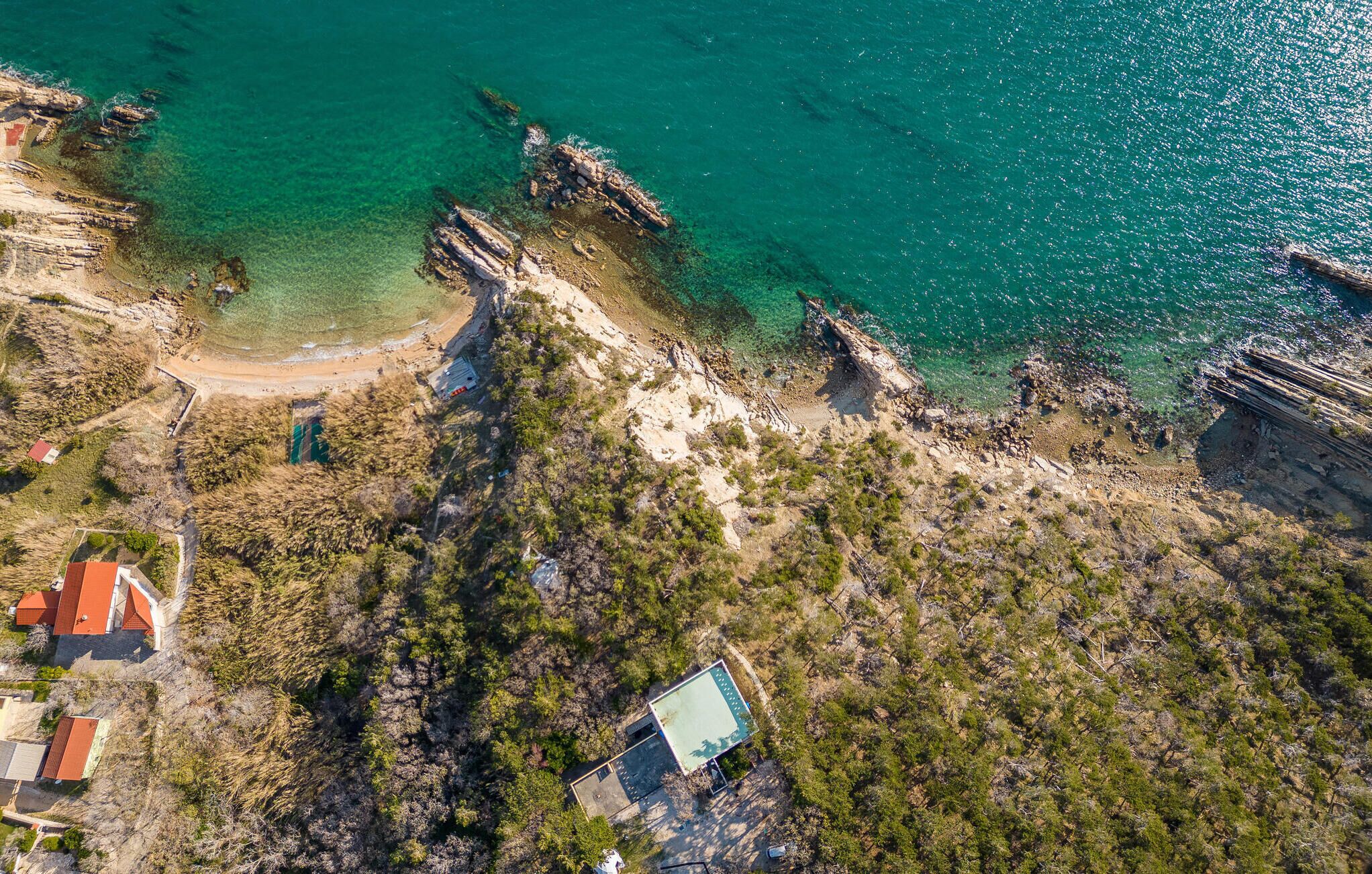 An aerial view of a coastal property with a beach, rocky cliffs, and a few buildings.