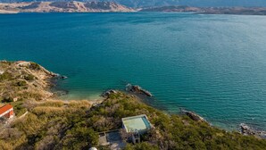 A coastal area with a clear view of the sea and mountains in the background. There is a small building with a red roof near the water's edge. - Cozy apartment in Novalja with WiFi (Novalja)