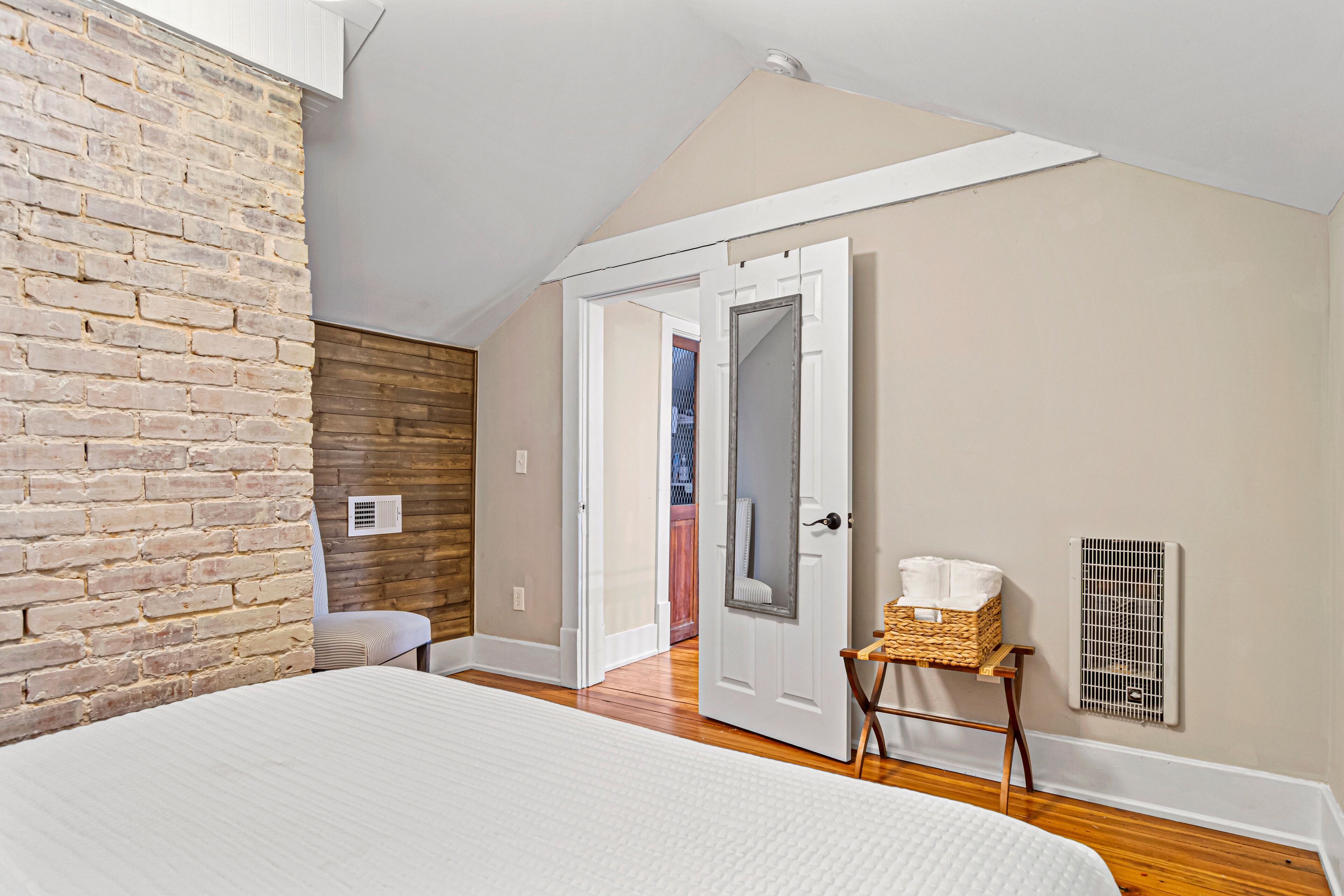 Un dormitorio con pared de ladrillo, suelo de madera, una cama, una puerta, una mesita con una cesta y un radiador.