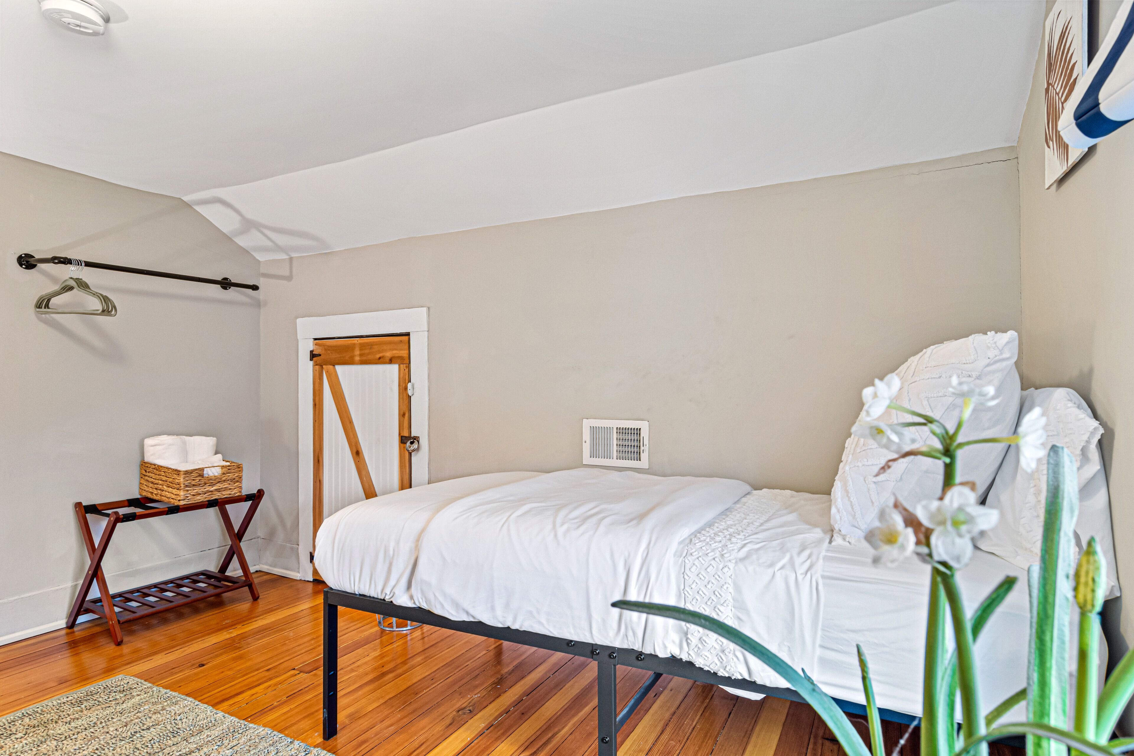 A bedroom with a bed, wooden floor, a small table with a basket, and a hanging rack.